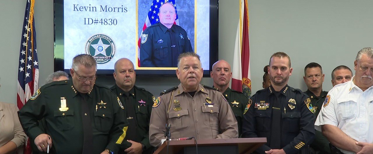 Sheriff Wayne Ivey announces the death of Senior Homicide Agent Kevin Morris, whose memorial portrait is displayed behind him.
